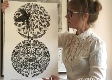 Artist standing with linocut print of a parrot, frogs and insects in a sand timer. 